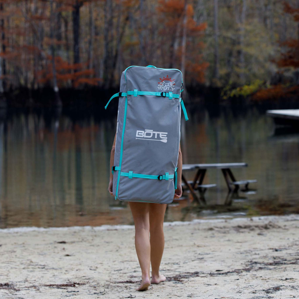 Person carries the WULF Aero 11′4″ Native Dune Inflatable Paddle Board in a bag on their back near the lake.