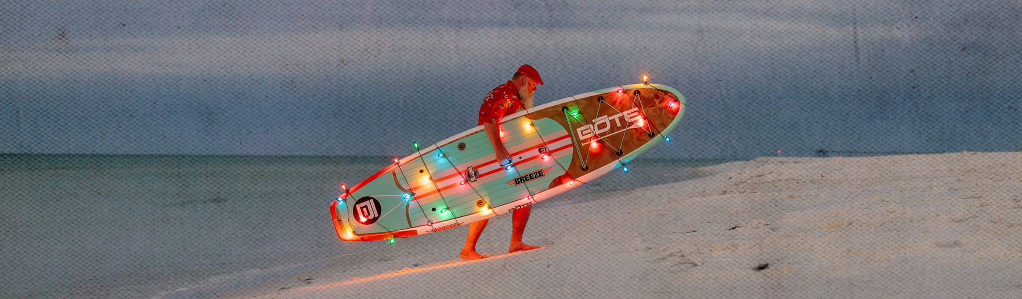 Person in red outfit standing on the beach holding a paddle board with colorful lights