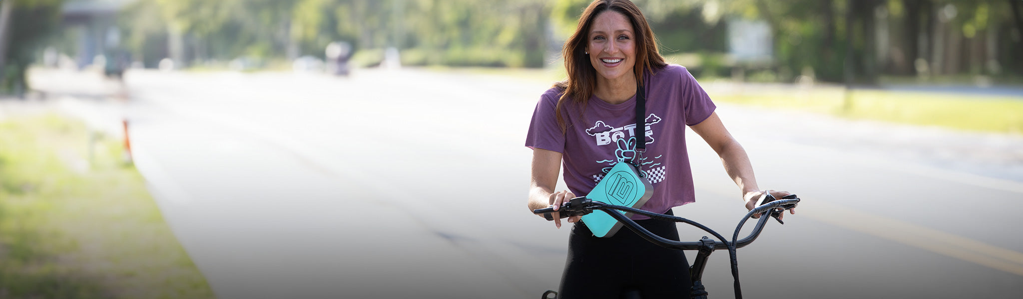 Smiling woman riding a bike with a turquoise crossbody case on a sunny street.