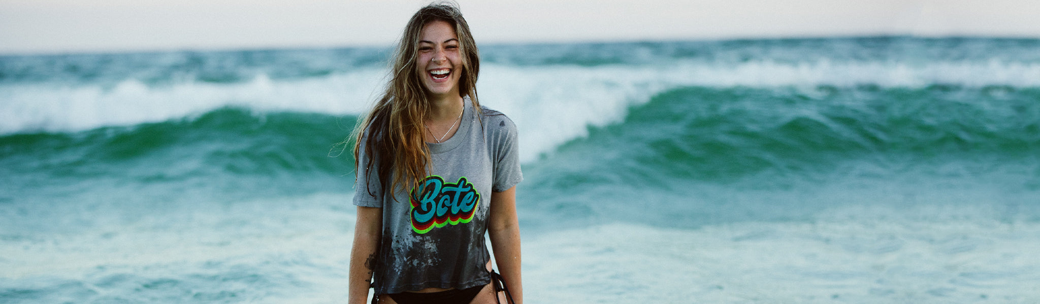 Woman smiling in wet gray logo tee standing in ocean waves near the shore.