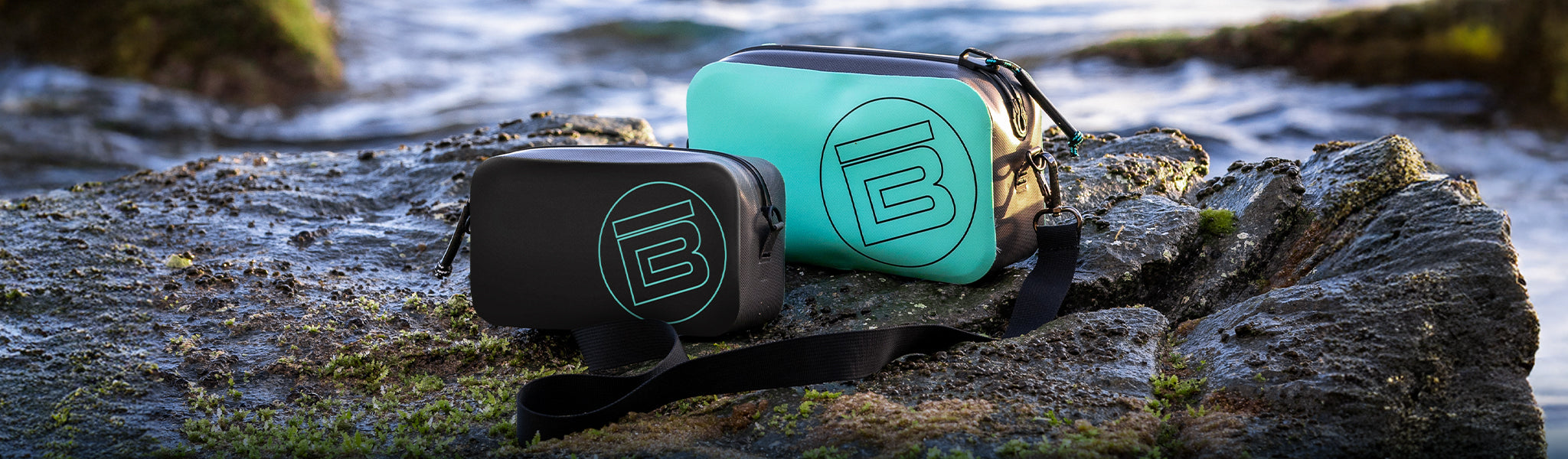 Black and turquoise waterproof bags resting on mossy rocks near ocean waves.