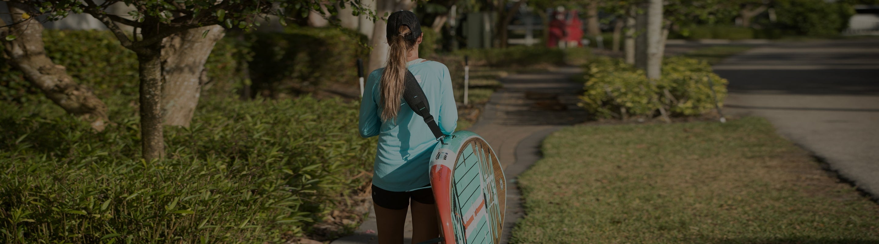 Woman walking down shaded path carrying teal paddle board under her arm.