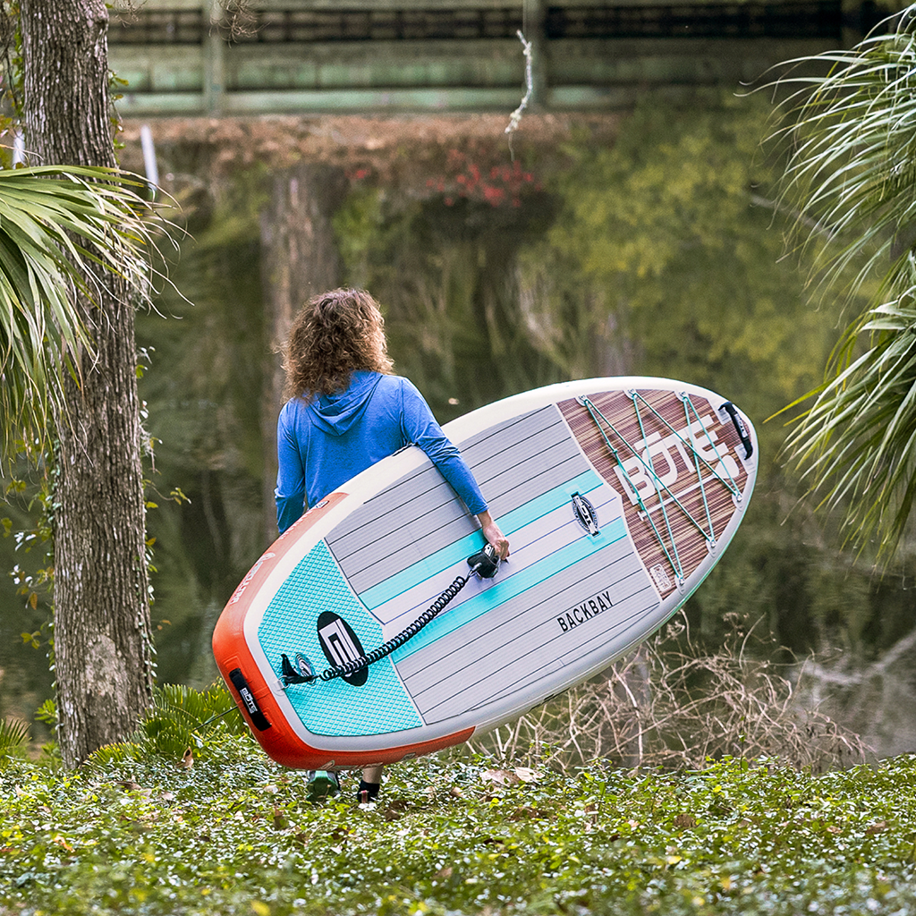 Person carrying Backbay Aero Inflatable Paddle Board toward a calm river, with teal deck pad and leash attached.