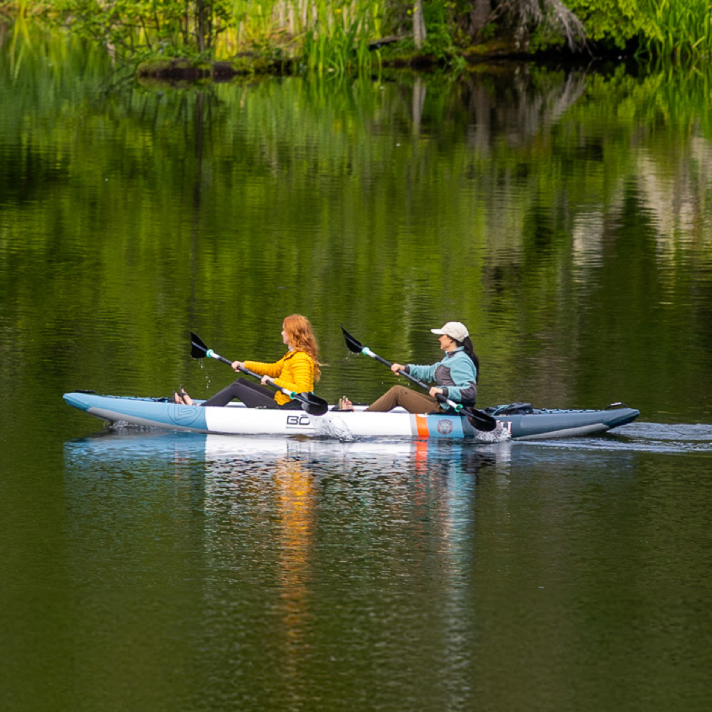 Two women paddling the Vali Aero kayak on a still green lake surrounded by trees.