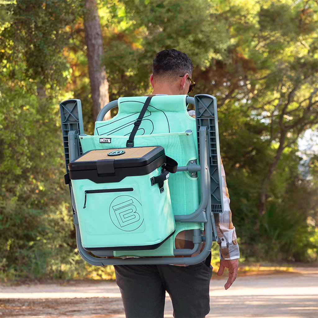 Person carrying folded chair with attached KULA Mule cooler outdoors.