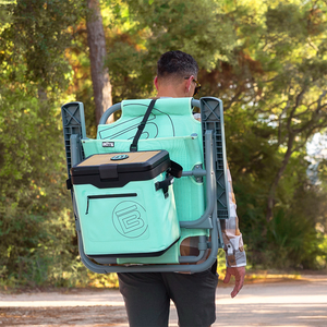 Person carrying folded chair with attached KULA Mule cooler outdoors.